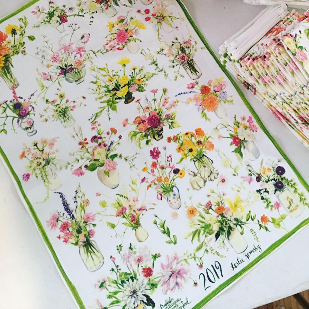 Artist, Leslie Zemsky's 2019 floral design printed on our tea towels. One towel is spread on the table and folded ones are next to it.