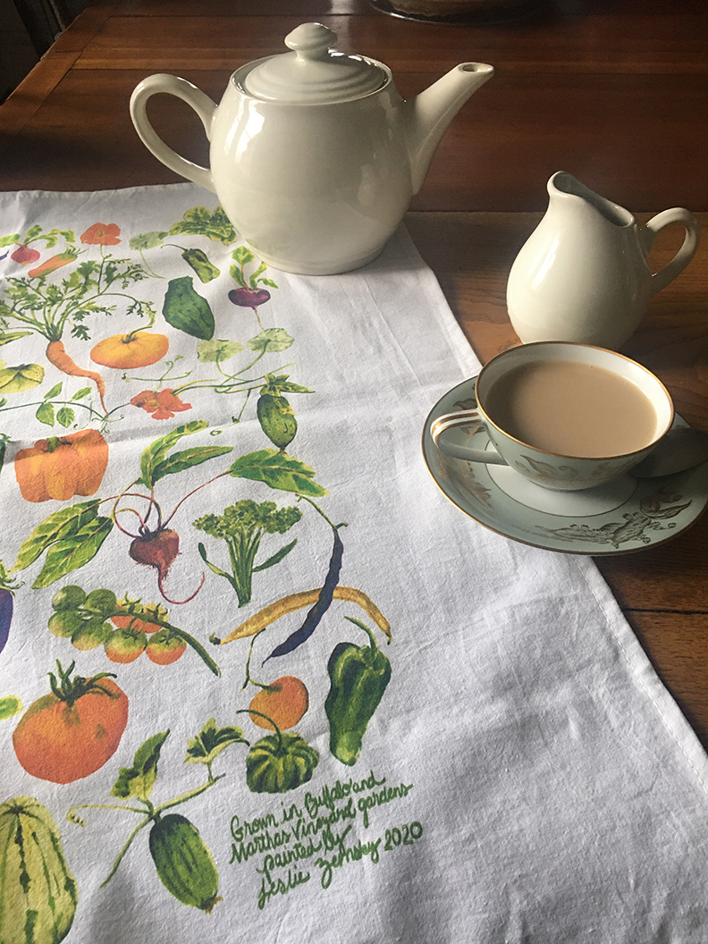 Artist Leslie Zemsky Veggie collage printed onto tea towel. It is spread on the table with a tea pot, tea cup filled with tea and a creamer arrangment