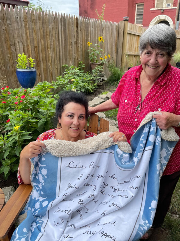 Mother and daughter sitting in a garden holding the Heirloom blanket with the mother's note to her daughter printed on it. The blanket is made of exceptionally soft faux mink with a sherpa back lining.