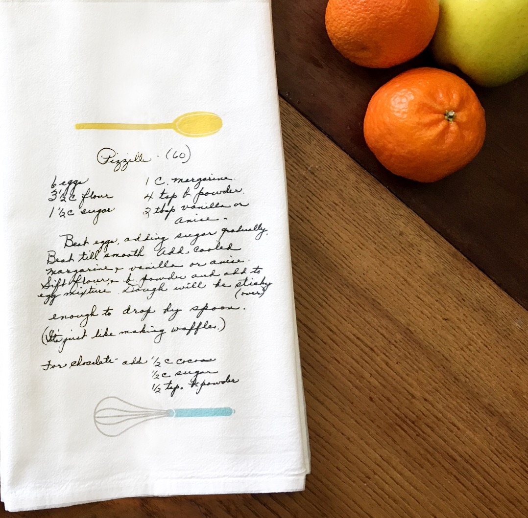 Heirloom Recipe Tea Towel with a handwritten recipe printed on it. There's a whisk and spoon design embelishments added. It sits on a table with 2 tangerines and an apple in the corner of the photo.